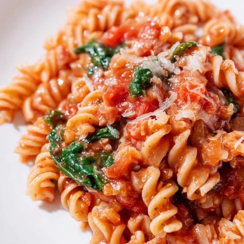 A close-up of Tomato Spinach One-Pot Rotini in a skillet, with wilted greens and diced tomatoes in a savory broth.  