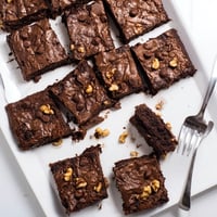 Golden-brown, crackly top brownies with a fudgy interior are stacked on a white plate, ready to be shared as a decadent dessert.