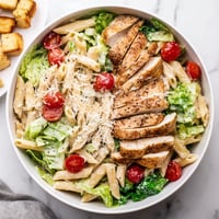 Tender grilled chicken slices rest atop a colorful Caesar pasta bowl with crisp romaine and cherry tomatoes, ready to serve.