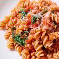 A close-up of Tomato Spinach One-Pot Rotini in a skillet, with wilted greens and diced tomatoes in a savory broth.  